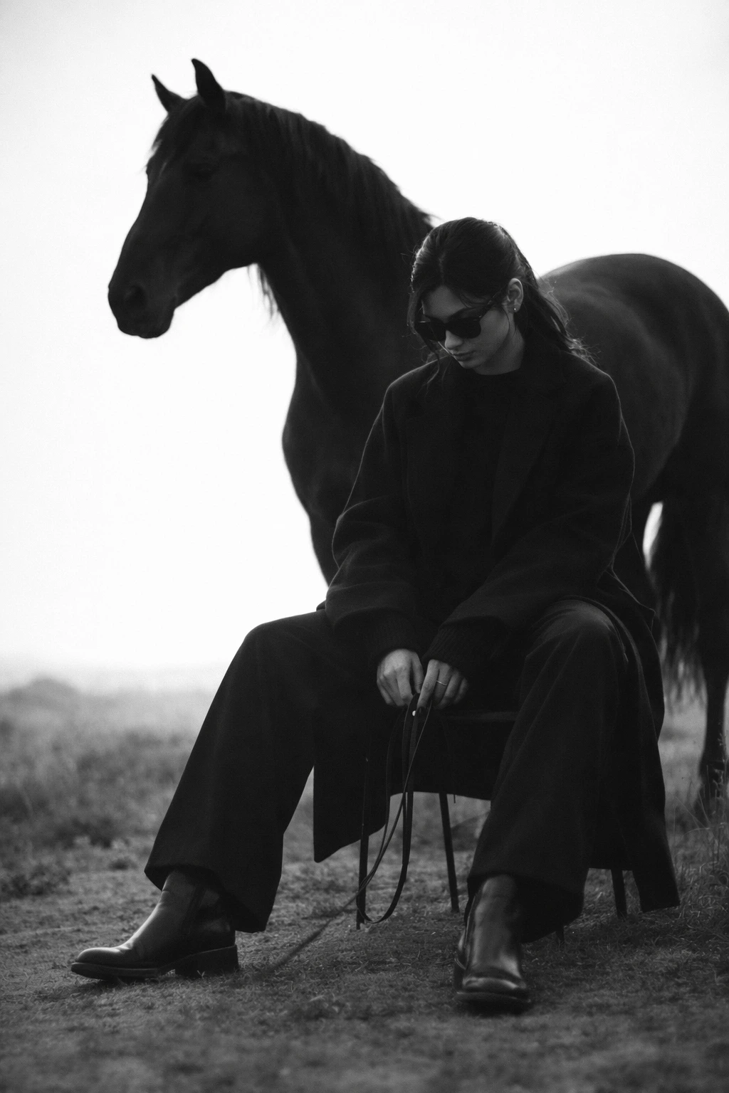 Cinematic black-and-white fashion editorial showing a seated figure in oversized dark layers and leather boots, holding reins while a black horse stands closely behind in an open landscape under an overexposed sky.